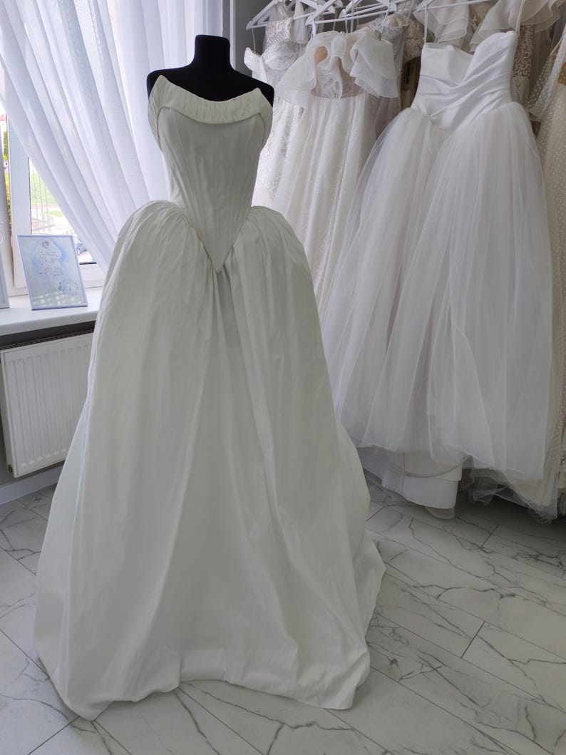 May include: A white wedding dress on a black mannequin. The dress has a fitted bodice and a full skirt. Other white wedding dresses hang in the background. The setting is a bridal shop or atelier.