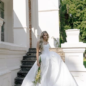 May include: A white wedding dress with a full skirt and a simple bodice. The bride is holding a bouquet of yellow flowers. The photo is taken outdoors in front of a white building with stone steps.