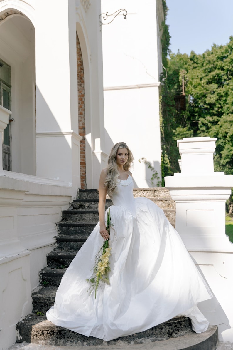 May include: A white wedding dress with a full skirt and a simple bodice. The bride is holding a bouquet of yellow flowers. The photo is taken outdoors in front of a white building with stone steps.