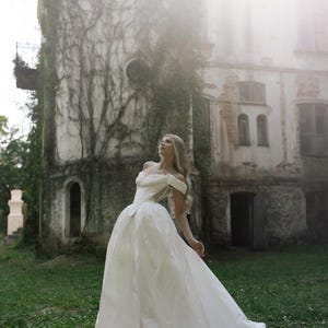 May include: A woman in a white off-the-shoulder wedding gown stands in a grassy area in front of a weathered stone building covered in vines. The dress has a full skirt and a long train, creating a romantic and ethereal scene.