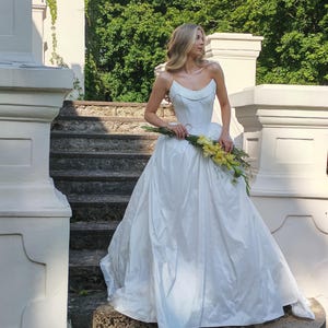 May include: A strapless white wedding gown with a full skirt. The bride is holding a bouquet of yellow flowers. The photo is taken outdoors on a stone staircase, with white pillars and green foliage in the background.