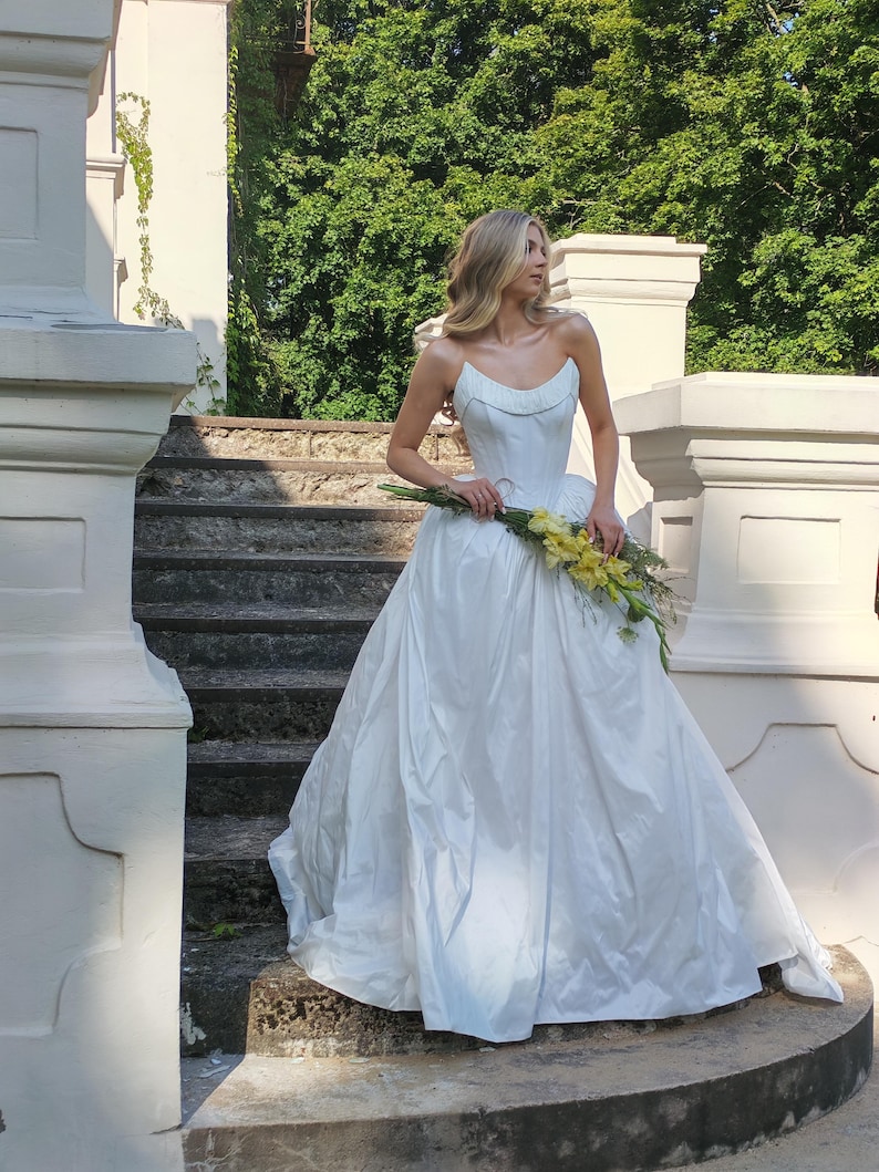 May include: A strapless white wedding gown with a full skirt. The bride is holding a bouquet of yellow flowers. The photo is taken outdoors on a stone staircase, with white pillars and green foliage in the background.