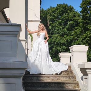 May include: A white strapless wedding gown with a full skirt and long train. The bride is standing on a stone staircase in front of a white building with green trees in the background. The dress has a fitted bodice.