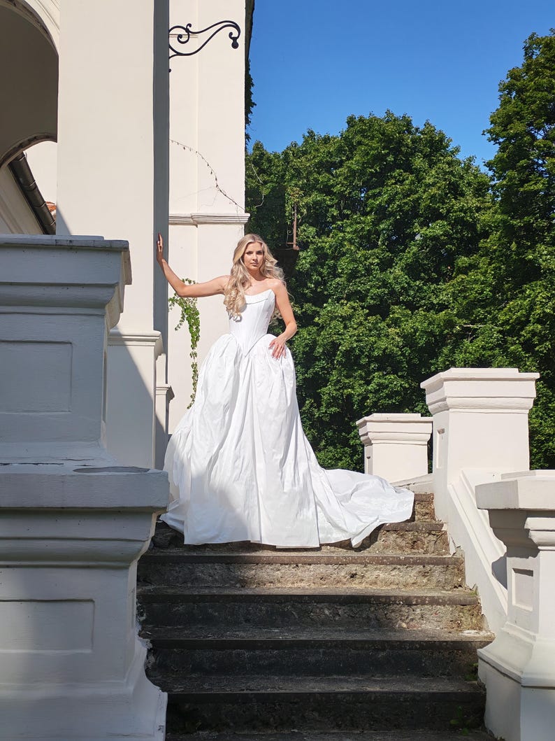 May include: A white strapless wedding gown with a full skirt and long train. The bride is standing on a stone staircase in front of a white building with green trees in the background. The dress has a fitted bodice.