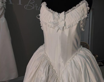 May include: A white wedding dress on a black mannequin. The dress features a sweetheart neckline with lace trim, a fitted bodice, and a full skirt. A small bow adorns the bodice. A pearl necklace is visible.