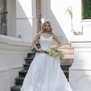 May include: A strapless white wedding dress with a full skirt. The bride holds a bouquet of yellow flowers. The dress has a fitted bodice and a flowing skirt. The background features a white building with stairs.
