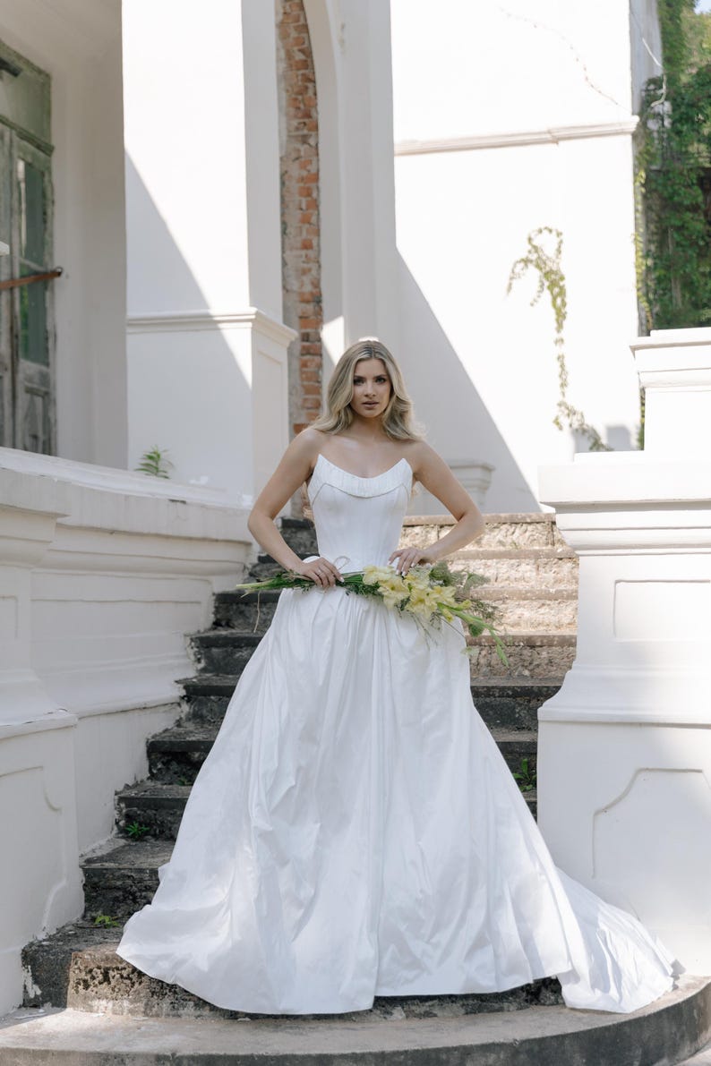 May include: A strapless white wedding dress with a full skirt. The bride holds a bouquet of yellow flowers. The dress has a fitted bodice and a flowing skirt. The background features a white building with stairs.