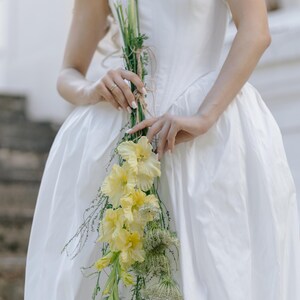 May include: A strapless white dress with a full skirt. A bouquet of yellow gladiolus flowers and greenery is held in front of the dress. The flowers are tied with twine. The background is a blurred outdoor setting.