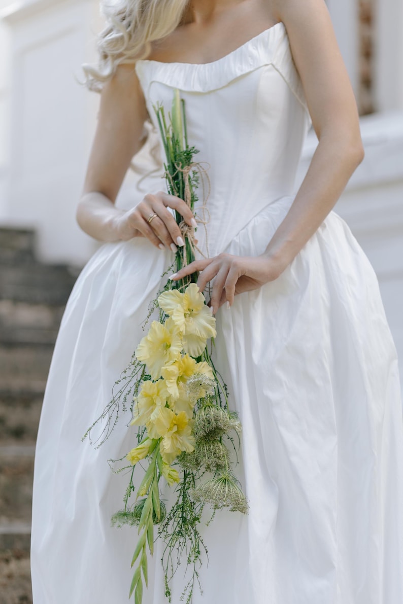 May include: A strapless white dress with a full skirt. A bouquet of yellow gladiolus flowers and greenery is held in front of the dress. The flowers are tied with twine. The background is a blurred outdoor setting.