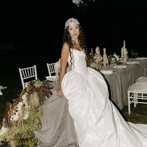 May include: A white wedding dress with a full skirt and a long train. The dress is paired with a lace headpiece. The scene is set outdoors at night, with a table set for a formal dinner, decorated with flowers and candles.