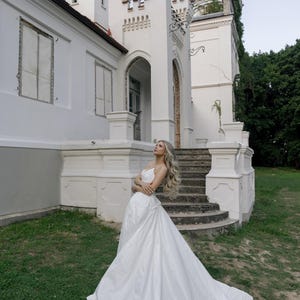 May include: A woman in a white wedding dress poses in front of a white castle-like building with a stone staircase. The dress has a long train, and the woman has long, wavy blonde hair. The building has decorative turrets.