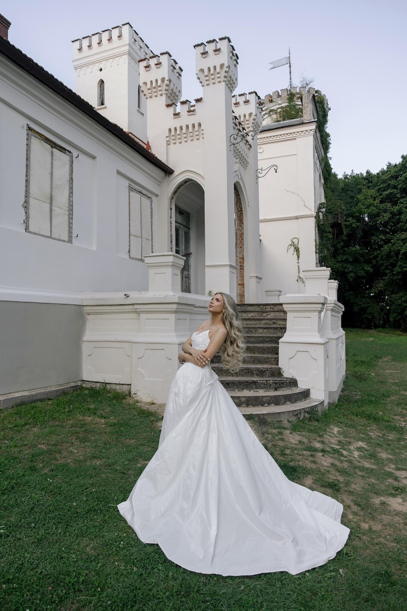 May include: A woman in a white wedding dress poses in front of a white castle-like building with a stone staircase. The dress has a long train, and the woman has long, wavy blonde hair. The building has decorative turrets.