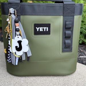 May include: An olive green YETI tote bag with black detailing, a black handle, and a decorative charm. The charm features a white flower with the letter "J", beads, and ribbons. The YETI logo is on a black patch.