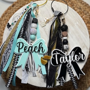 Könnte beinhalten: Zwei personalisierte Schlüsselanhänger mit den Namen "Peach" und "Taylor". Jeder Schlüsselanhänger hat einen charmförmigen Anhänger, bunte Bänder, Perlen und einen Metallverschluss. Die Schlüsselanhänger befinden sich auf einer weißen Holzoberfläche.