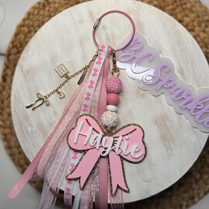 May include: A pink and gold keychain with a bow-shaped charm that says "Haytie". The keychain has a pink ring, ribbons, and decorative charms. The background is a white wooden circle.