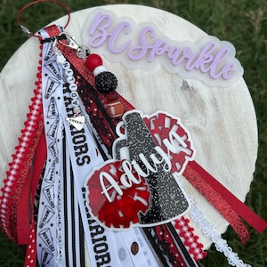 May include: A personalised keychain with red, black, and white ribbons, beads, and charms. The keychain features the name "Adilyn" and the text "BC Sparkle". Decorative elements include a football charm and cheerleader pom-pom accents.