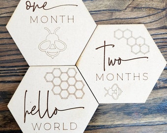 May include: Three wooden hexagonal baby milestone signs with a bee and honeycomb design. The signs read "one month", "two months", and "hello world".