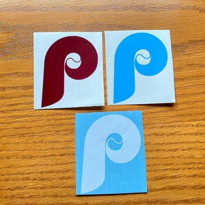May include: Three stickers featuring the Philadelphia Phillies logo. The logos are in red, blue, and white. The stickers are on white paper.
