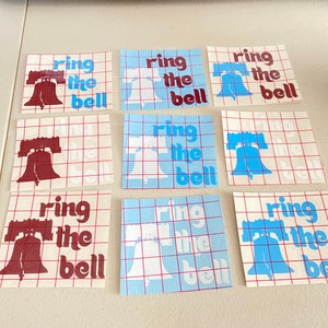 May include: Nine sheets of transfer paper with a grid pattern. Each sheet has a design of the Liberty Bell with the text "ring the bell" in a retro font. Three sheets have a maroon bell and text, three sheets have a white bell and text, and three sheets have a blue bell and text.