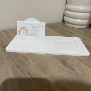 May include: White acrylic and wood display stand with a small, white, round tray. A business card with the text "RILA CO. Acrylic and wood keepsakes" is displayed on the stand.