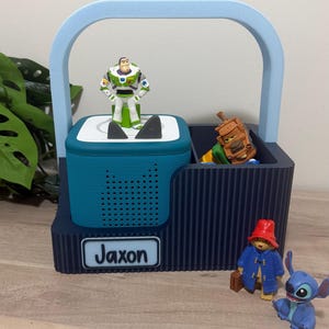 May include: A dark blue 3D-printed storage caddy with a light blue handle, featuring a teal speaker dock with a Buzz Lightyear figure on top. The caddy has a white nameplate reading Jaxon, with a brown truck, a red-hatted bear, and a blue alien figure.
