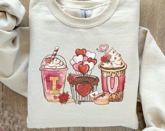 Puede incluir: Sudadera de color crema con un diseño del Día de San Valentín. El diseño incluye tres bebidas ilustradas: una bebida helada rosa con un corazón, una magdalena con globos de corazón y una bebida de café con una pajita en forma de corazón. También hay rosas rojas y corazones.