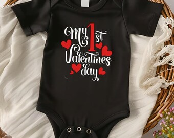 May include: Black baby bodysuit with the text "My 1st Valentines day" in white script and red hearts. The number "1" is in red. The bodysuit has short sleeves and three snaps at the bottom.