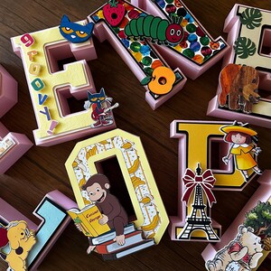 May include: A collection of three-dimensional letters in pastel pink, yellow, and light blue, decorated with various children's book characters and motifs including a caterpillar, a monkey reading a book, a dog with balloons, and an Eiffel Tower.