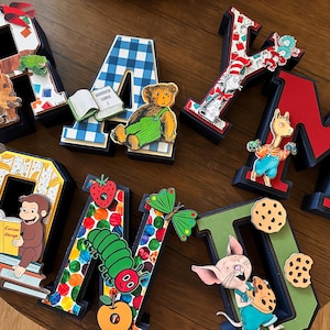 May include: A set of decorative 3D letters spelling RAYMOND, each featuring unique children's book character illustrations including a brown bear, a teddy bear, Curious George, a caterpillar, and a piglet, with colorful patterns and motifs.
