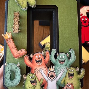 May include: A large, three-dimensional letter O in olive green with a black border, decorated with cutouts of monsters, a child in a white wolf costume, and letters spelling WILD. The design features vibrant, cartoon-style illustrations of creatures.