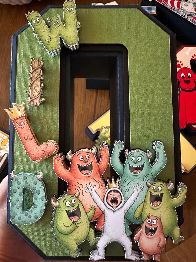 May include: A large, three-dimensional letter O in olive green with a black border, decorated with cutouts of monsters, a child in a white wolf costume, and letters spelling WILD. The design features vibrant, cartoon-style illustrations of creatures.