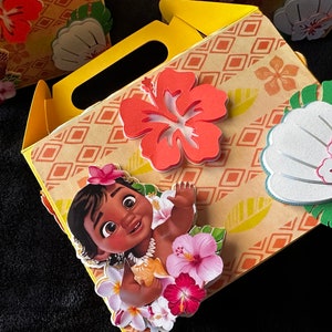 May include: A yellow cardboard party favor box with a Moana character on the front. The box is decorated with flowers and leaves.