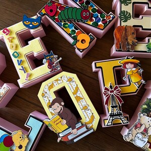 May include: A collection of three-dimensional block letters in various colors, decorated with cutouts of children's book characters including a caterpillar, a monkey reading a book, a bear, and an Eiffel Tower, with the word GROOVY printed on one letter.