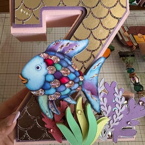 May include: A three-dimensional number seven decoration featuring a gold scale pattern, a blue and purple fish with iridescent scales, and layered paper cutouts of green and purple seaweed. A ruler shows the base measures 7 inches wide.