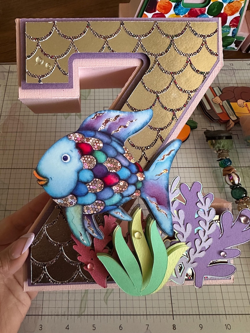 May include: A three-dimensional number seven decoration featuring a gold scale pattern, a blue and purple fish with iridescent scales, and layered paper cutouts of green and purple seaweed. A ruler shows the base measures 7 inches wide.