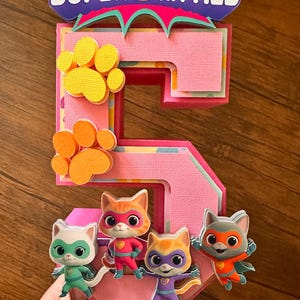 Puede incluir: Decoración de cumpleaños con el número cinco rosa y en capas con las palabras "SUPER KITTIES" en la parte superior. El número tiene huellas de patas amarillas y naranjas y cuatro figuras de gatos de dibujos animados con disfraces de superhéroes.