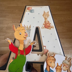 May include: A large, three-dimensional letter A featuring a light-colored background with blue polka dots and illustrations of toys. A cartoon llama in green overalls is attached to the left, and a circular image of three llamas is on the right.