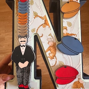 May include: A large three-dimensional letter N decorated with a pattern of monkeys in trees. A figure in a black suit and patterned trousers stands on the left, balancing a tall stack of colorful flat caps. Three additional flat caps are attached.