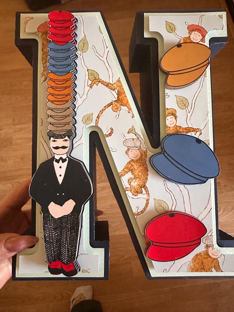 May include: A large three-dimensional letter N decorated with a pattern of monkeys in trees. A figure in a black suit and patterned trousers stands on the left, balancing a tall stack of colorful flat caps. Three additional flat caps are attached.