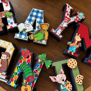 May include: A collection of seven three-dimensional letters spelling RAYMOND, each decorated with colorful children's book illustrations including a brown bear, a blue gingham pattern, a teddy bear, Cat in the Hat, Curious George, a caterpillar, and Piglet.