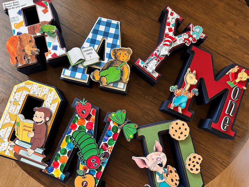 May include: A collection of seven three-dimensional letters spelling RAYMOND, each decorated with colorful children's book illustrations including a brown bear, a blue gingham pattern, a teddy bear, Cat in the Hat, Curious George, a caterpillar, and Piglet.