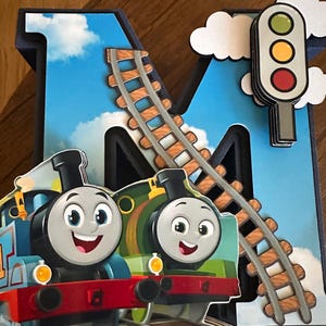 May include: A large, three-dimensional letter M decorated with a blue sky and white clouds background. It features a curved train track, a traffic light with green, yellow, and red circles, and two cartoon train engines in blue and green.