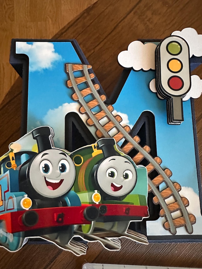 May include: A large, three-dimensional letter M decorated with a blue sky and white clouds background. It features a curved train track, a traffic light with green, yellow, and red circles, and two cartoon train engines in blue and green.