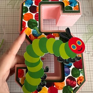 May include: A three-dimensional letter S crafted from light pink cardstock, featuring a colorful pattern of overlapping red, orange, yellow, green, and blue circles. A layered paper cutout of a green caterpillar with a red head is attached.