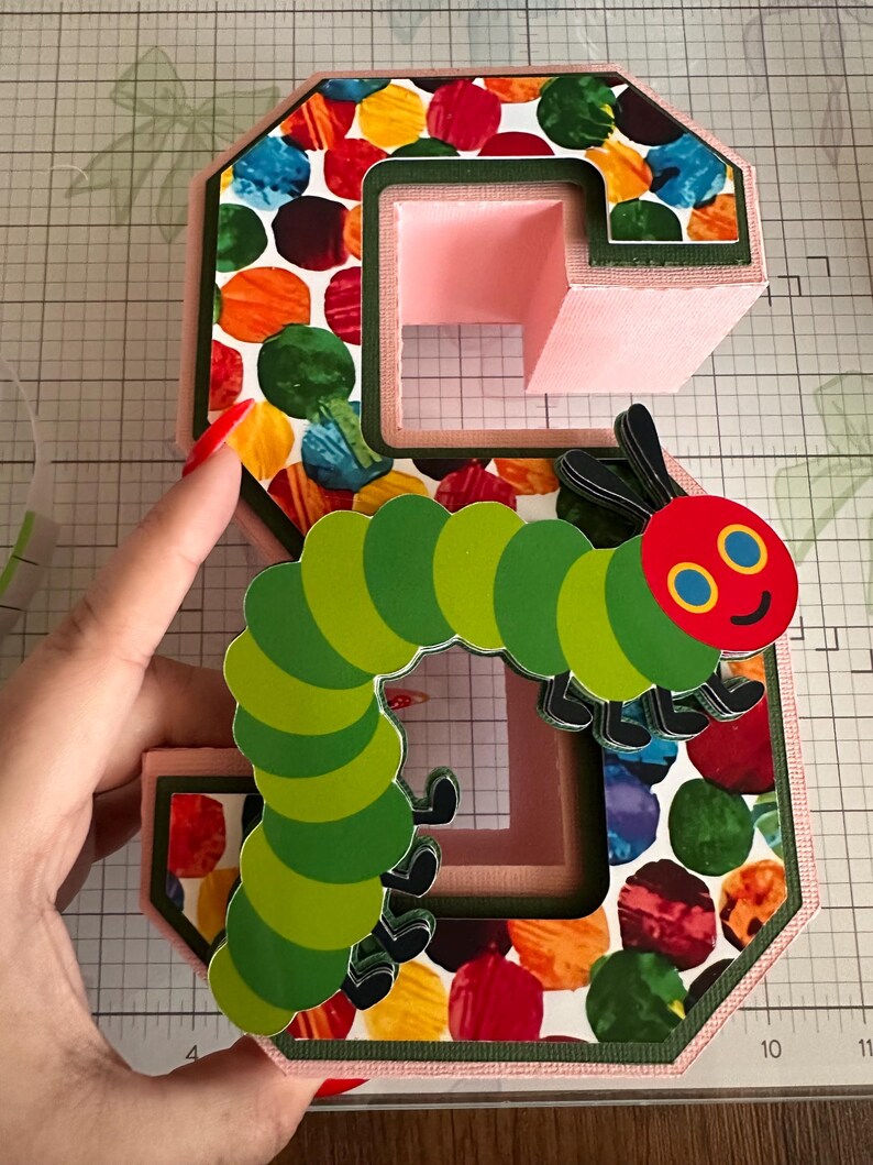 May include: A three-dimensional letter S crafted from light pink cardstock, featuring a colorful pattern of overlapping red, orange, yellow, green, and blue circles. A layered paper cutout of a green caterpillar with a red head is attached.