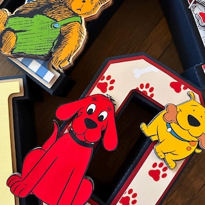 May include: A decorative letter O featuring a cream background with red paw prints and white bone patterns. A bright red dog character is attached to the left side, and a yellow dog character holding a bone is attached to the right side.