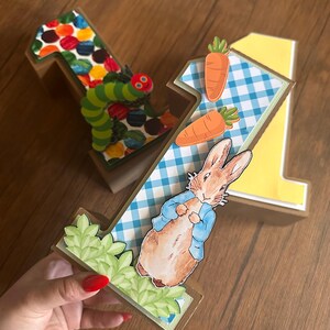 May include: Two three-dimensional number one decorations. One features a colorful dot pattern and a green caterpillar. The other features a blue and white gingham pattern, two orange carrots, and a brown rabbit wearing a blue jacket with green leaves.
