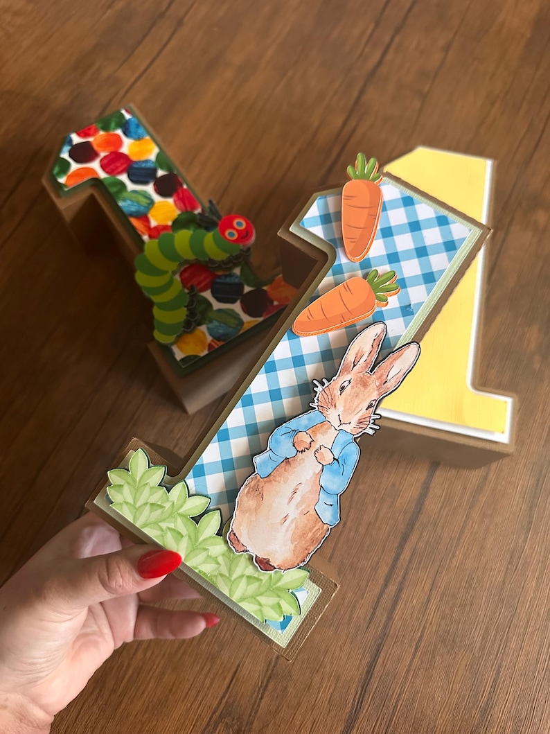 May include: Two three-dimensional number one decorations. One features a colorful dot pattern and a green caterpillar. The other features a blue and white gingham pattern, two orange carrots, and a brown rabbit wearing a blue jacket with green leaves.