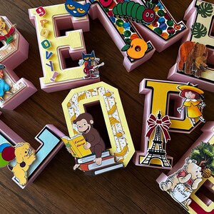 May include: A set of three-dimensional letters spelling PENELOPE, each decorated with colorful children's book characters and motifs including Pete the Cat, The Very Hungry Caterpillar, Curious George, Madeline, and Winnie the Pooh.