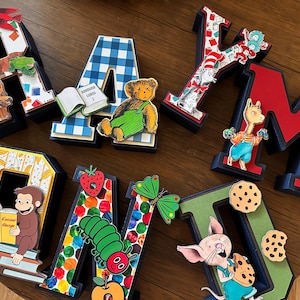 May include: A set of eight three-dimensional letters spelling RAYMOND, decorated with various children's book characters and motifs including a brown bear, a blue plaid pattern, a cat in a hat, a monkey, a caterpillar, and a pig eating cookies.